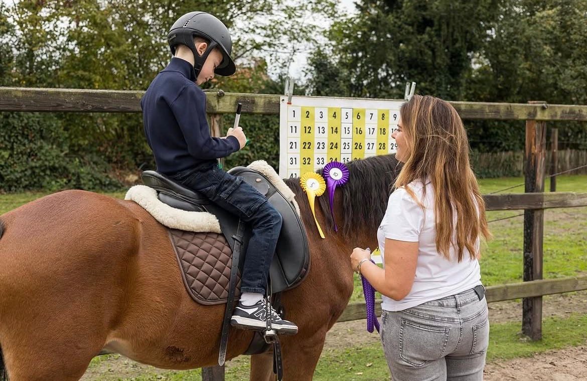 Kind op bruin paard met helm tijdens bijles met cijferbord en paarse strik