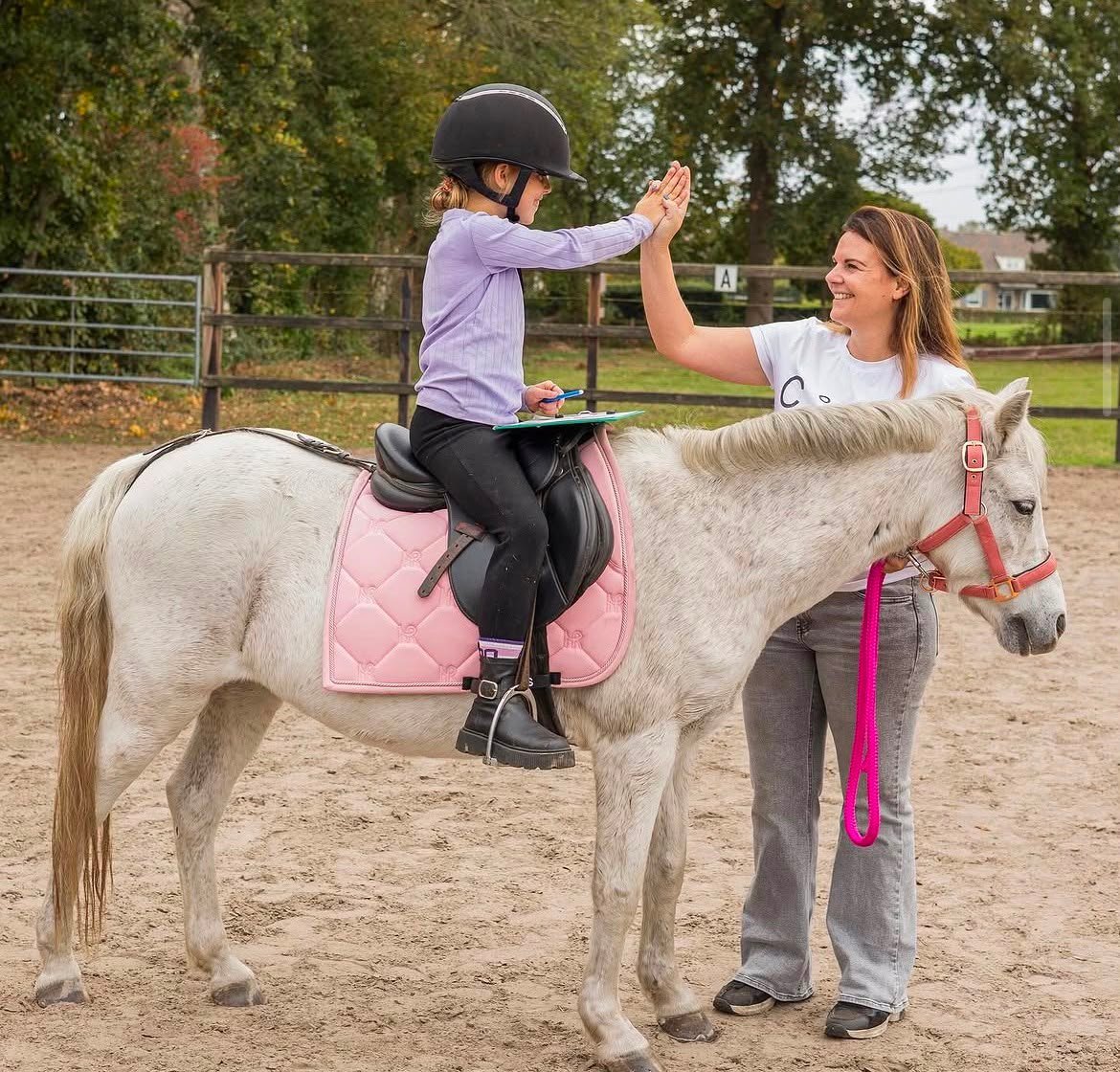Jong kind op witte pony met helm geeft high five aan begeleidster die de roze leidtouw vasthoudt in de bak