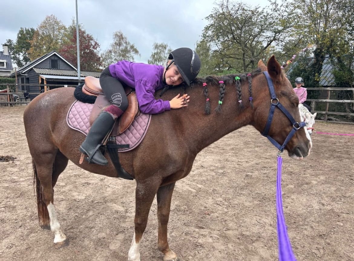 Kind in paars jack ligt ontspannen op een bruine pony met zadel in de buitenbak